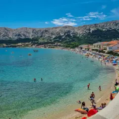 Słoneczna plaża na wyspie Krk tętni życiem. Ludzie kąpią się w krystalicznie czystej wodzie, opalają pod parasolami i spacerują brzegiem.