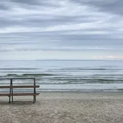 Pusta ławka na plaży, morze i chmury. W oddali widać żaglówkę.