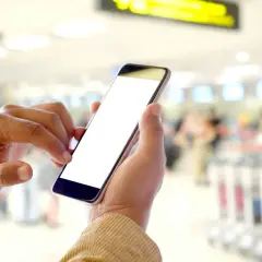 Ręce trzymające smartfon z pustym ekranem, pokazujące, na czym polega odprawa online w tle lotniska.