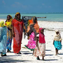 Kobiety i dziewczynki w kolorowych strojach spacerują po plaży Zanzibaru. Myślą o wakacjach i o tym, jakie **szczepienia** są potrzebne.