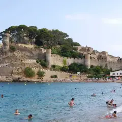 Ludzie kąpią się w morzu przy plaży z zamkiem na wzgórzu. Zastanawiasz się, ile się leci do Hiszpanii, by zobaczyć takie widoki?