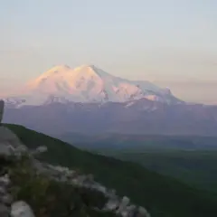 Ośnieżony, najwyższy szczyt Kaukazu, Elbrus, lśni w blasku zachodzącego słońca. Przed nim zielone wzgórza i skaliste zbocze.