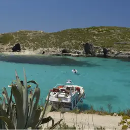 Malta: turkusowa woda, łodzie i skaliste wybrzeże. Ludzie spacerują, kąpią się. Czy na Malcie jest bezpiecznie? Tak, to raj dla turystów.