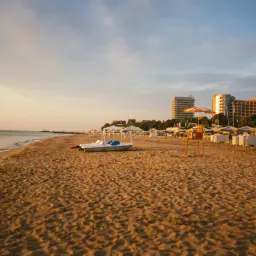 Złote piaski plaży o zachodzie słońca, z łódką, leżakami i hotelami w tle.