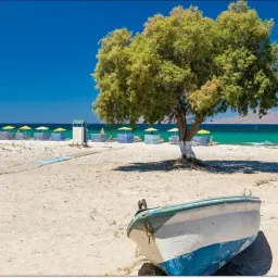 Piękna plaża na wyspie Kos. Łódka na piasku, drzewo, leżaki i parasole pod błękitnym niebem.