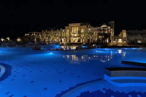 Nocny basen z niebieskim podświetleniem i luksusowym hotelem Lazuli w tle.
