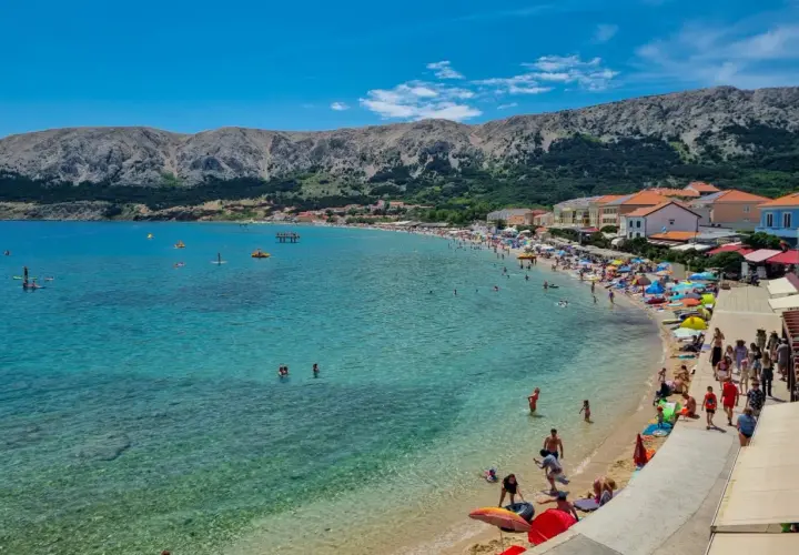 Słoneczna plaża na wyspie Krk tętni życiem. Ludzie kąpią się w krystalicznie czystej wodzie, opalają pod parasolami i spacerują brzegiem.