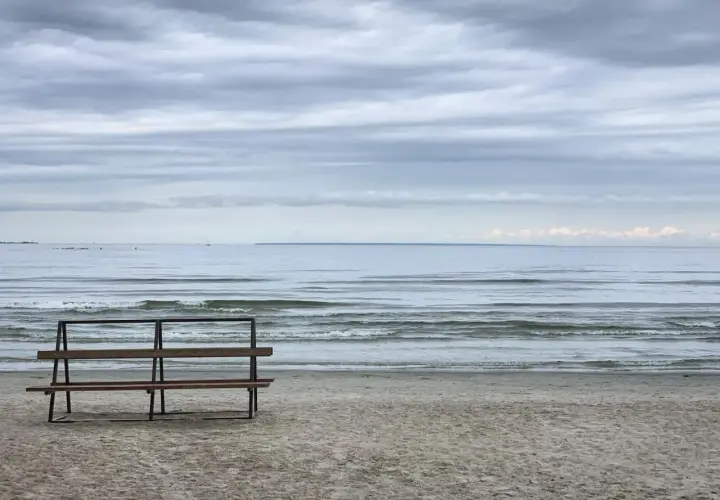 Pusta ławka na plaży, morze i chmury. W oddali widać żaglówkę.