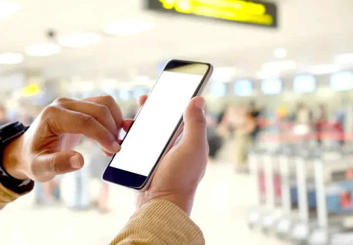 Ręce trzymające smartfon z pustym ekranem, pokazujące, na czym polega odprawa online w tle lotniska.