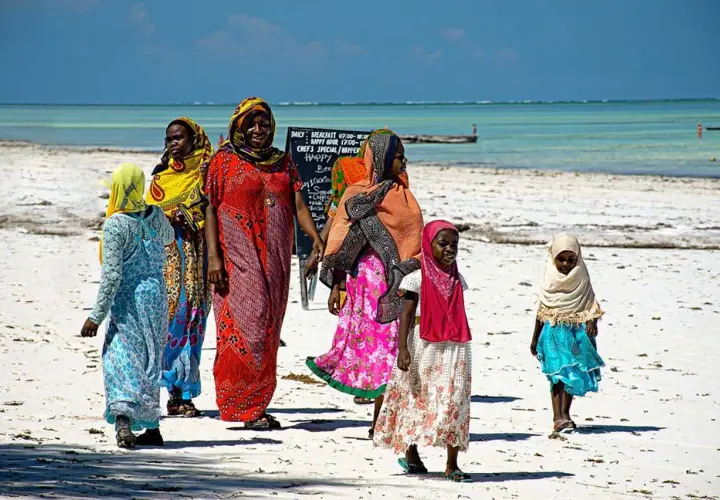 Kobiety i dziewczynki w kolorowych strojach spacerują po plaży Zanzibaru. Myślą o wakacjach i o tym, jakie **szczepienia** są potrzebne.