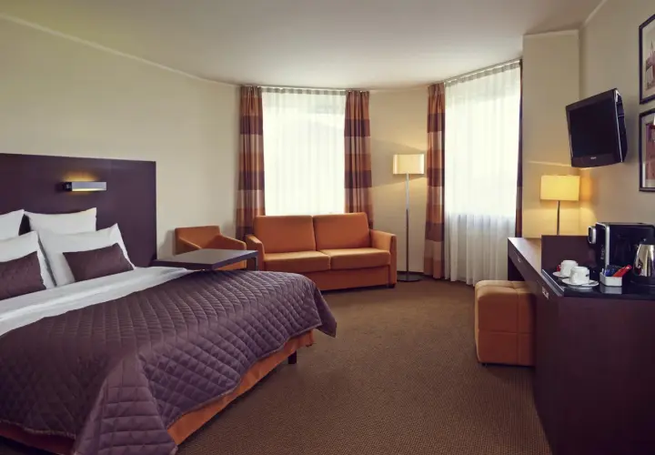 Przytulny pokój w hotelu Mercure Warszawa Airport: łóżko, kanapa, biurko z ekspresem do kawy i TV.