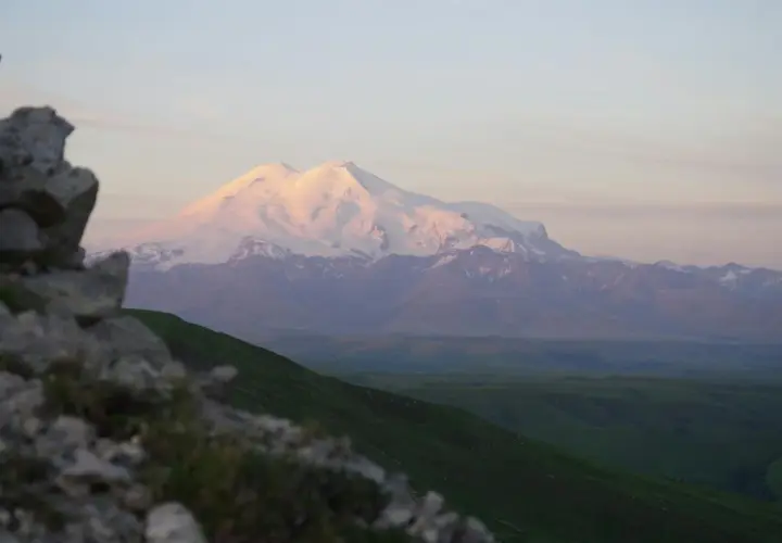 Ośnieżony, najwyższy szczyt Kaukazu, Elbrus, lśni w blasku zachodzącego słońca. Przed nim zielone wzgórza i skaliste zbocze.