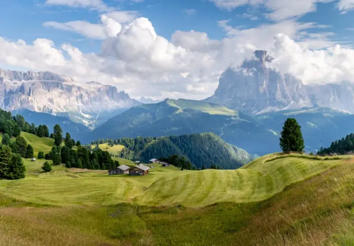 Zielone wzgórza i drewniane chatki w Val Gardena, z majestatycznymi górami w tle.