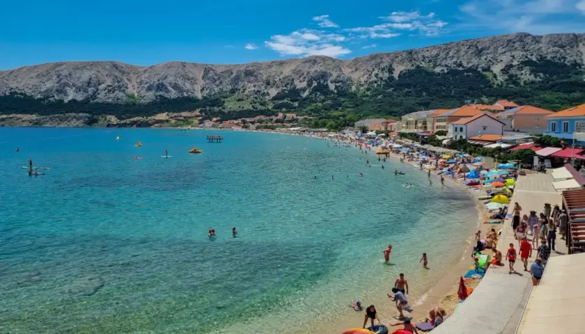 Słoneczna plaża na wyspie Krk tętni życiem. Ludzie kąpią się w krystalicznie czystej wodzie, opalają pod parasolami i spacerują brzegiem.