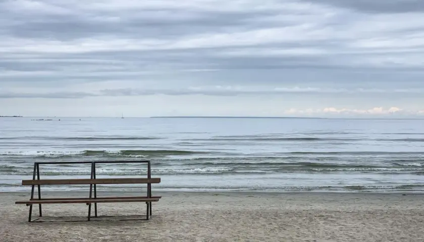 Pusta ławka na plaży, morze i chmury. W oddali widać żaglówkę.