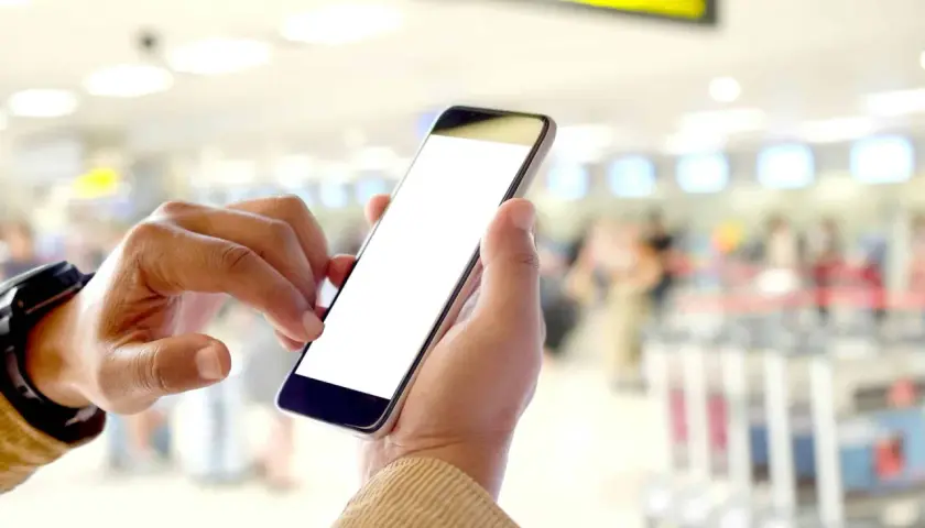 Ręce trzymające smartfon z pustym ekranem, pokazujące, na czym polega odprawa online w tle lotniska.