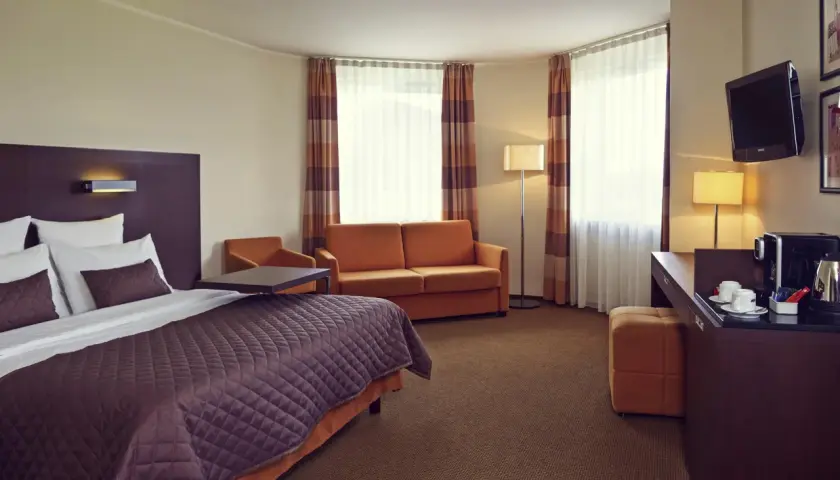 Przytulny pokój w hotelu Mercure Warszawa Airport: łóżko, kanapa, biurko z ekspresem do kawy i TV.