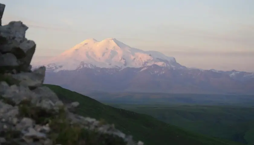 Ośnieżony, najwyższy szczyt Kaukazu, Elbrus, lśni w blasku zachodzącego słońca. Przed nim zielone wzgórza i skaliste zbocze.