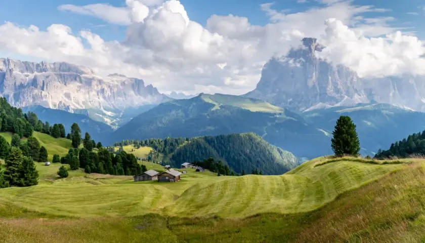 Zielone wzgórza i drewniane chatki w Val Gardena, z majestatycznymi górami w tle.