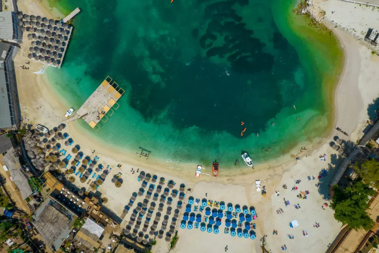 Albania, idealne miejsce na wakacje! Widok z lotu ptaka na turkusową wodę, piaszczystą plażę z leżakami i parasolami, drewniane molo i łodzie.