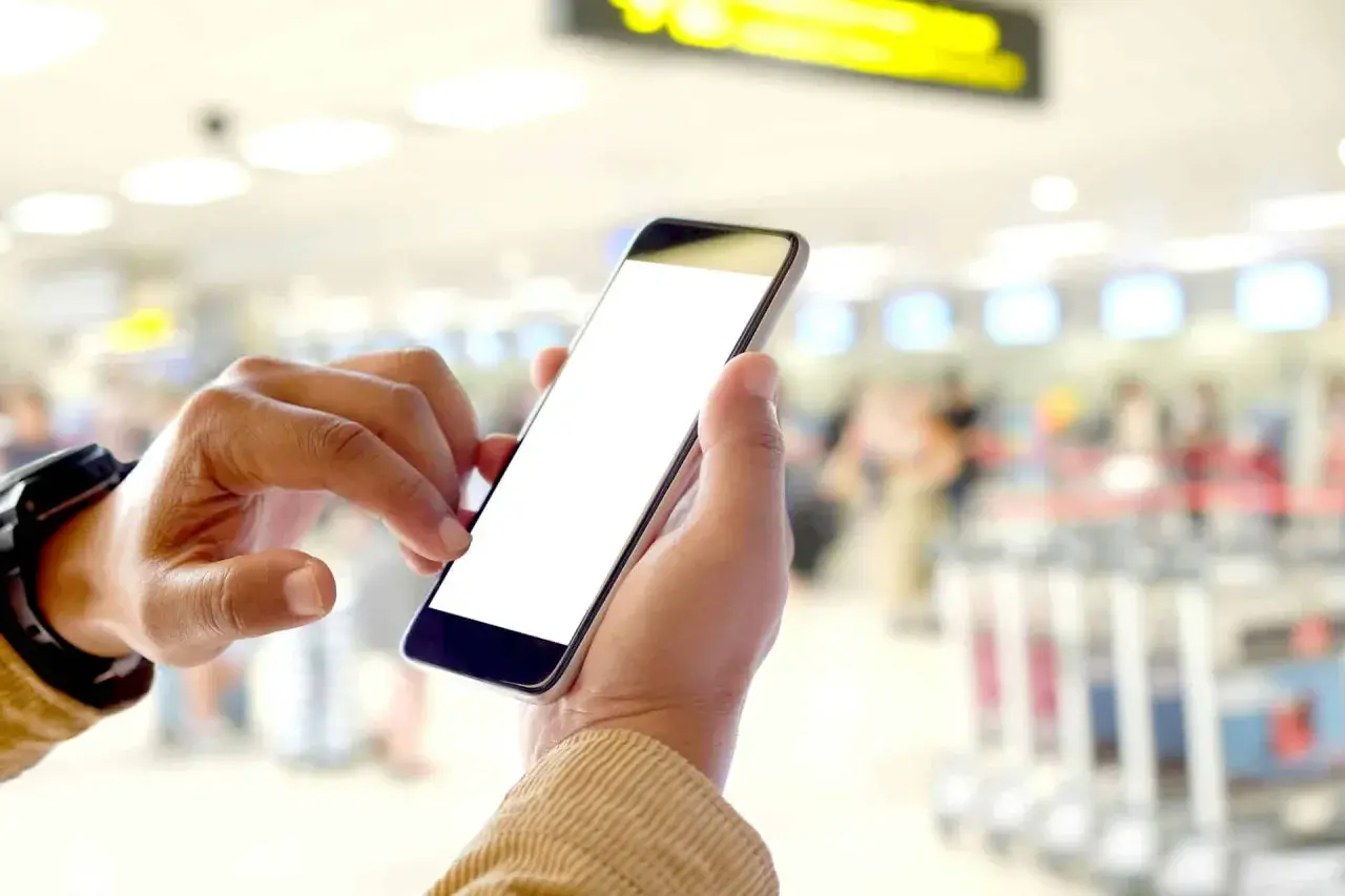 Ręce trzymające smartfon z pustym ekranem, pokazujące, na czym polega odprawa online w tle lotniska.