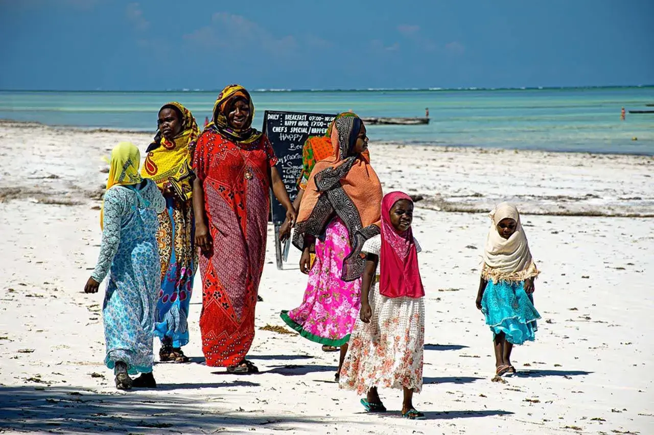 Kobiety i dziewczynki w kolorowych strojach spacerują po plaży Zanzibaru. Myślą o wakacjach i o tym, jakie **szczepienia** są potrzebne.