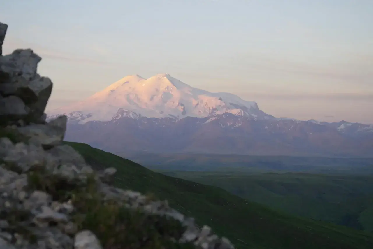 Ośnieżony, najwyższy szczyt Kaukazu, Elbrus, lśni w blasku zachodzącego słońca. Przed nim zielone wzgórza i skaliste zbocze.