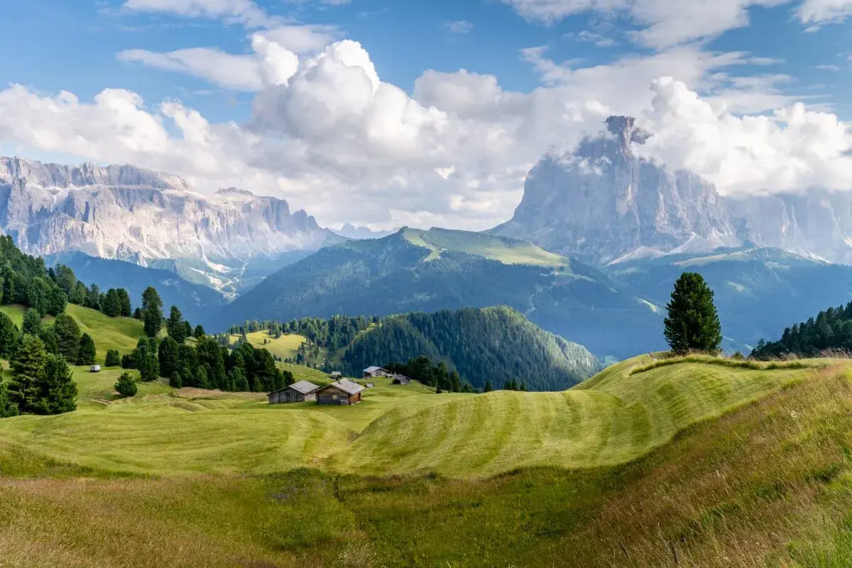 Zielone wzgórza i drewniane chatki w Val Gardena, z majestatycznymi górami w tle.