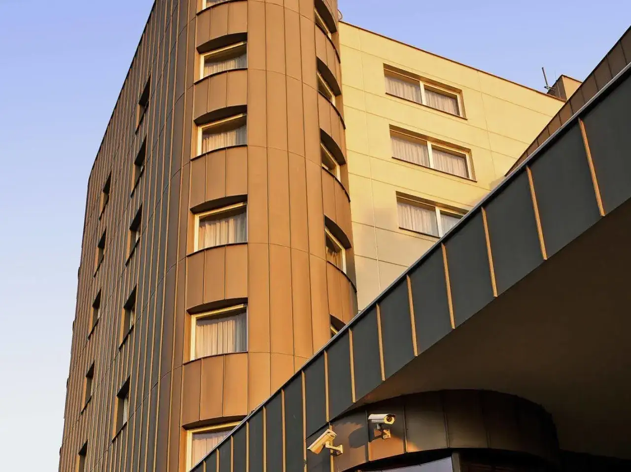 Hotel Mercure Warszawa Airport z nowoczesną, zaokrągloną fasadą i oknami w ciepłym świetle zachodzącego słońca.