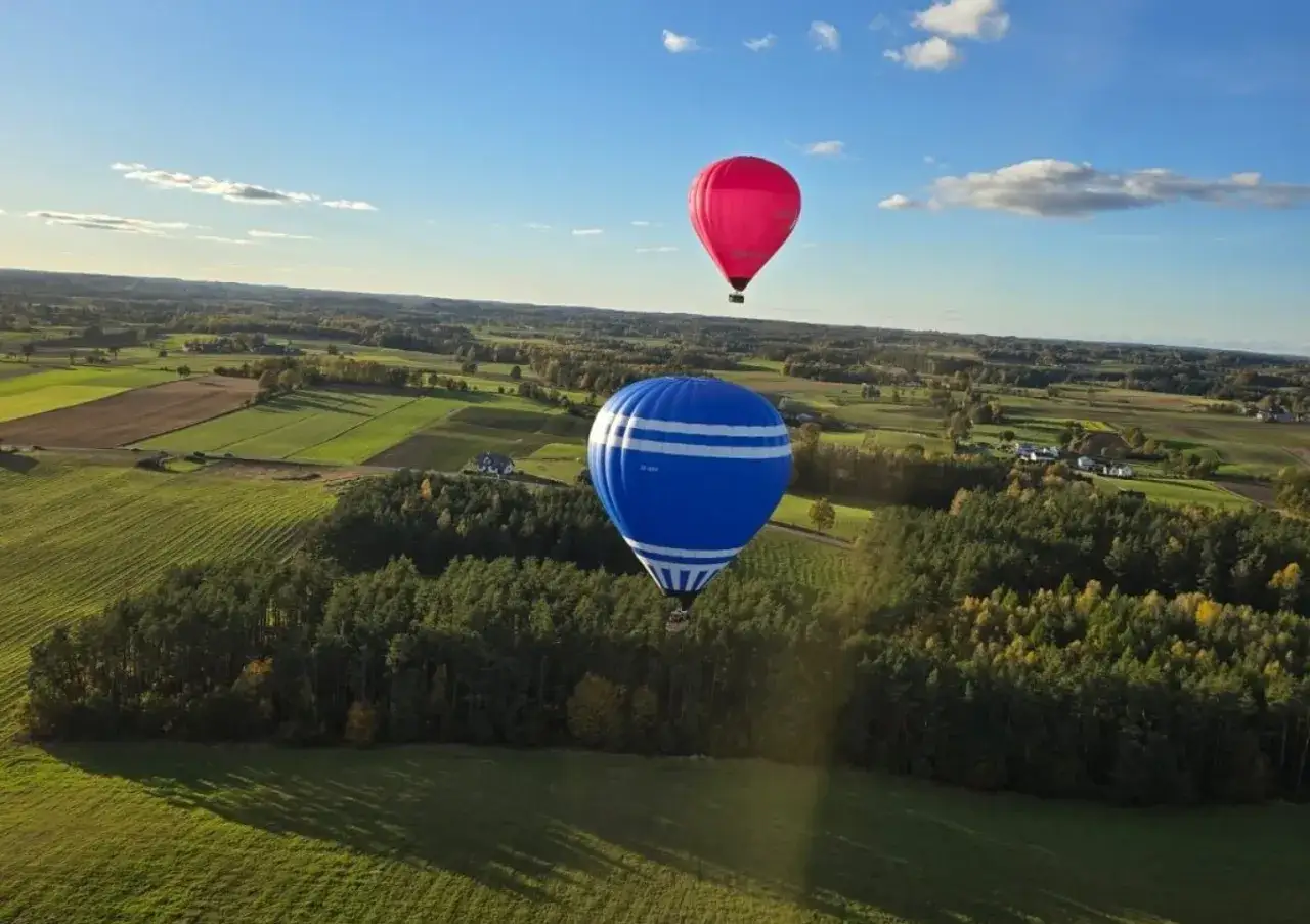 Dwa balony, jeden niebieski w białe pasy, drugi różowy, szybują nad polami i lasami. Niezapomniany lot balonem Poznań.
