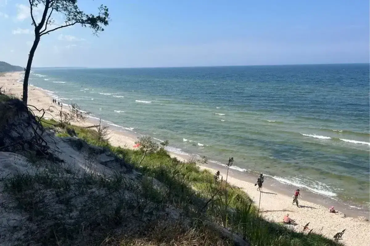 Widok na piaszczystą plażę i morze z wydmami porośniętymi trawą. W oddali widać spacerujących ludzi. Miejsce idealne na relaks, jak w orzechowo.