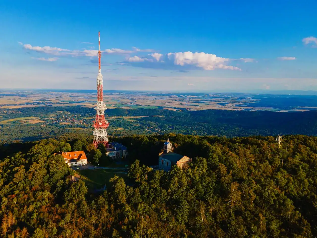 Wieża radiowa na szczycie góry Ślęża, otoczona lasem. W oddali rozciąga się krajobraz pól i wzgórz, a niebo jest błękitne z białymi chmurami. Góra Ślęża kryje wiele tajemnic.