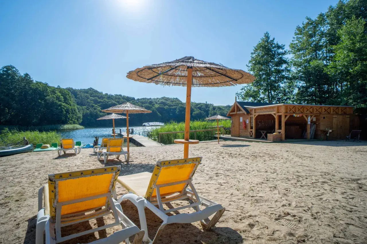 Plaża z leżakami i parasolami, idealne miejsce na wczasy dla samotnych po 60 tce. W tle jezioro i drewniana sauna.