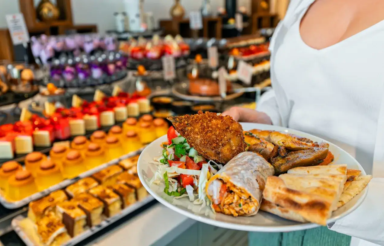 Talerz pełen smakołyków: panierowany kurczak, wrap, pieczone warzywa i pieczywo. W tle kuszący bufet słodkości i dań, prawdziwy full board plus.