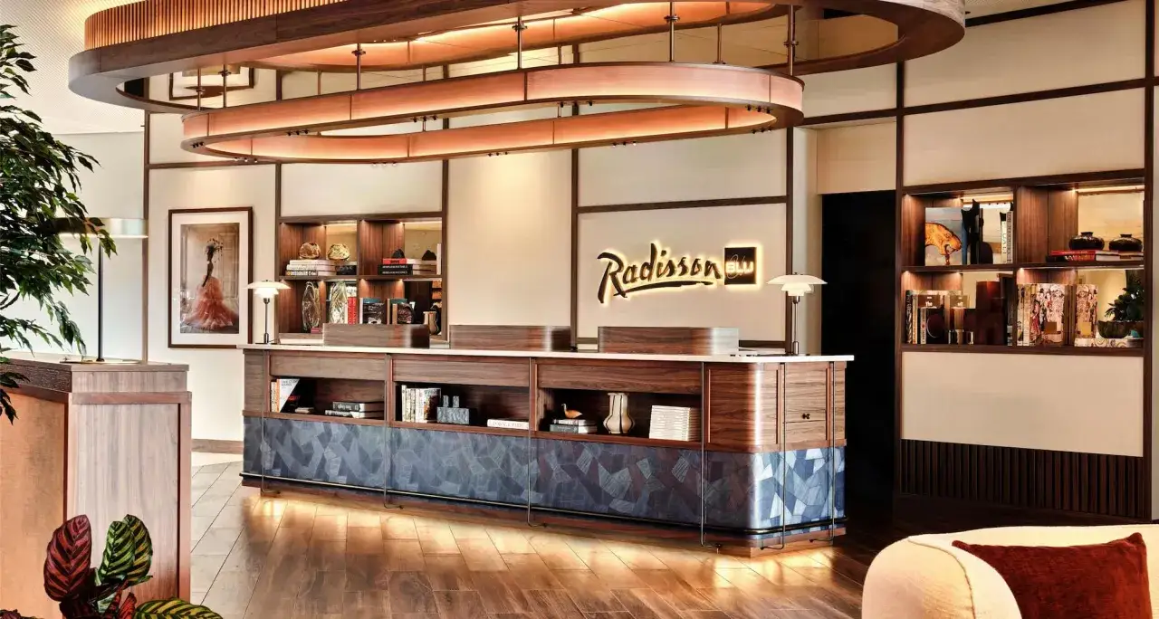 Eleganckie lobby hotelu Radisson Blu Airport Hamburg. Drewniana lada recepcyjna, ozdobne półki z książkami i dziełami sztuki, stylowe oświetlenie.
