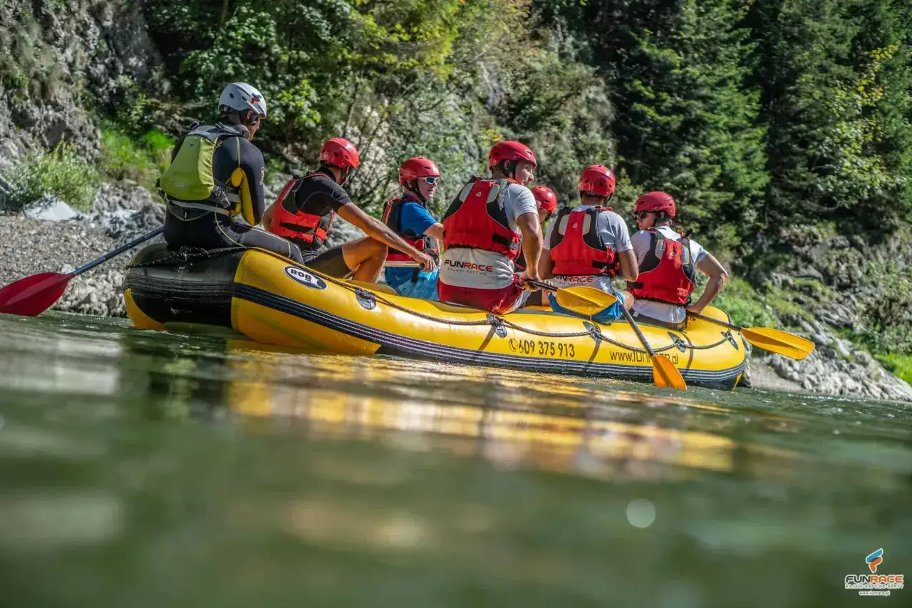Grupa osób w kaskach i kamizelkach na żółtej tratwie. To jest właśnie rafting – emocjonująca przygoda na rwącej rzece.