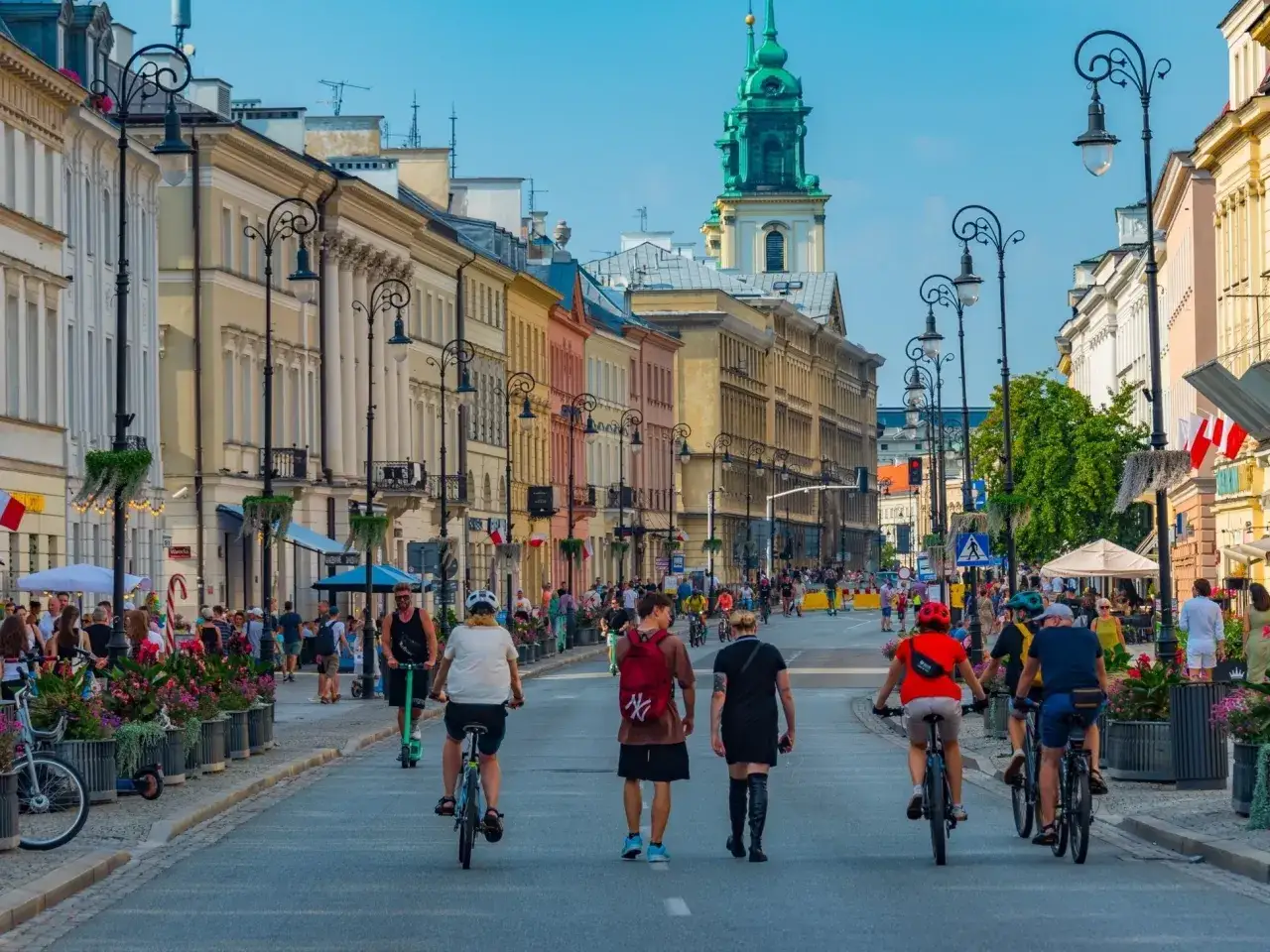 Tętniące życiem miasta w Polsce, gdzie ludzie jeżdżą na rowerach i hulajnogach po urokliwej ulicy z kolorowymi kamienicami i zieloną wieżą kościoła w tle.